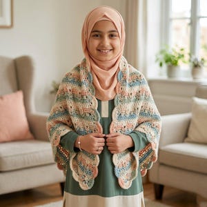 May include: A crocheted shawl in pastel colors of blue, peach, and cream. The shawl has a scalloped edge and is worn over a green dress. The person is wearing a peach-colored hijab.