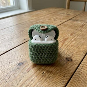 May include: A green crocheted case for wireless earbuds, secured with a wooden button. The case is open, displaying white earbuds. The case is on a wooden table.
