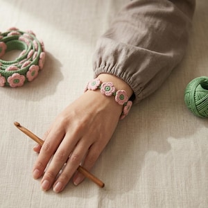 May include: A handmade crochet bracelet featuring pink flower-shaped designs with green centers and a green band. A matching crochet piece and a ball of green yarn are also visible. A wooden crochet hook rests on a light-colored surface.
