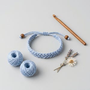 Könnte beinhalten: Ein hellblaues Häkelarmband mit Holzperlen, zwei Knäuel hellblauer Wolle und einem Häkelhaken aus Holz. Das Armband liegt neben einem kleinen Strauß Trockenblumen auf einer hellgrauen Oberfläche.