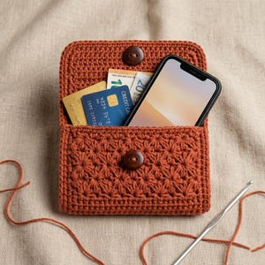Op de afbeelding: Een gehaakte, roestkleurige portemonnee met een sterrenpatroon. De portemonnee is open en toont een smartphone, creditcards en valuta. Het heeft een houten knoopsluiting. Een haaknaald en garen zijn zichtbaar.