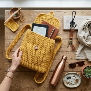 Könnte beinhalten: Ein senfgelber Häkelrucksack mit Knopfverschluss, der ein Tablet, ein Notizbuch und einen Stift enthält. Eine kupferfarbene Wasserflasche, Sonnenbrille und dekorative Anstecknadeln liegen auf einer Holzoberfläche.