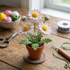 May include: A handcrafted crochet daisy flower arrangement in a small terracotta pot. The daisies have pink petals and yellow centers. The pot sits on a crocheted coaster. Other craft supplies, including yarn and scissors, are in the background.