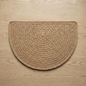 May include: A tan, semi-circular, crocheted rug. The rug features a concentric circular design in the center, with a border around the edge. The rug is made of a natural fiber and has a textured appearance. The rug is placed on a light-colored wooden surface.