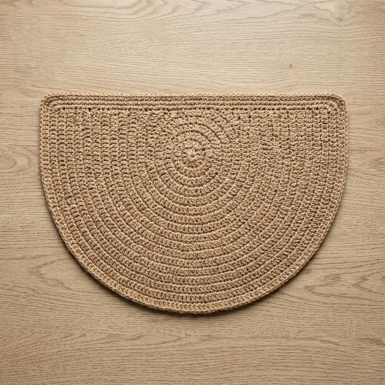 May include: A tan, semi-circular, crocheted rug. The rug features a concentric circular design in the center, with a border around the edge. The rug is made of a natural fiber and has a textured appearance. The rug is placed on a light-colored wooden surface.