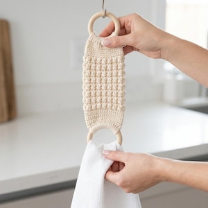 May include: A cream-colored, crocheted kitchen towel holder with wooden rings. The holder features a textured, bobble stitch design. A white waffle-weave towel is held in the bottom ring. The holder is suspended from a metal hook.