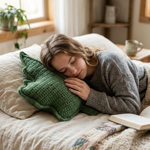 Pode incluir: Uma pessoa descansando em uma cama com uma almofada verde de crochê em forma de folha. A pessoa está vestindo um suéter cinza e coberta com um cobertor. Um livro e uma caneca são visíveis na cama.