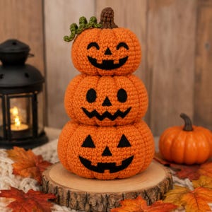 May include: A stacked crochet pumpkin decoration featuring three orange pumpkins with black felt jack-o'-lantern faces. The top pumpkin has a small green curly vine and a brown stem. The pumpkins are textured with yarn stitches and stacked vertically.