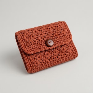 Puede incluir: Cartera de ganchillo de color terracota cálido. La cartera presenta un patrón de estrella y una solapa asegurada con un botón redondo de madera. La cartera hecha a mano es de tamaño compacto, ideal para llevar lo esencial.