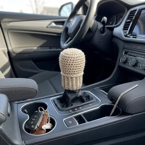 Pode incluir: Uma capa de manípulo de mudança de marcha bege de crochê é mostrada no interior de um carro. A capa é cilíndrica, com uma base canelada e um topo arredondado. O volante, o painel e a consola do carro são visíveis.