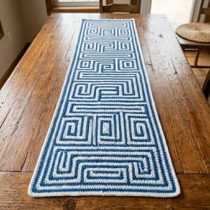 May include: A rectangular, blue and white crochet table runner with a geometric maze-like pattern. The runner is placed on a wooden table. The design features a repeating series of squares and lines, creating a visually interesting textile.
