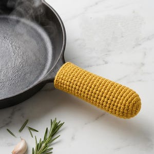 Puede incluir: Una sartén de hierro fundido negra con una funda de mango de ganchillo amarilla. Sale vapor de la sartén. Ramitas de romero y un diente de ajo están en primer plano, lo que sugiere cocinar.