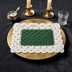 May include: A handmade, rectangular crochet doily with a white scalloped border and a green center, resting on a gold-colored plate. The image also includes a small silver salt cellar, two brass candle holders with lit candles, and a glass of water.
