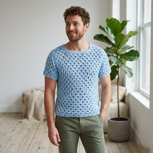 Pode incluir: Um top de crochê azul claro de manga curta com decote quadrado. O design de tecido aberto cria um padrão de grade. O top é usado com calças verde-oliva. A pessoa está em frente a uma janela com uma planta grande.