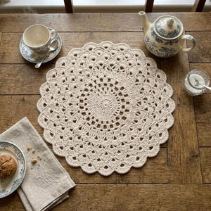 Puede incluir: Un tapete de ganchillo de color crema con un borde festoneado está sobre una mesa de madera. Una taza y un platillo, una tetera, un pequeño cuenco de azúcar y una servilleta con un plato y una galleta también están sobre la mesa. El tapete tiene aproximadamente 36 cm de diámetro.