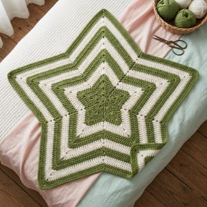 May include: A handmade, star-shaped crochet blanket in shades of green and white. The blanket features a five-pointed star design with alternating stripes of green and white. The blanket is displayed on a bed with a pink and blue sheet.