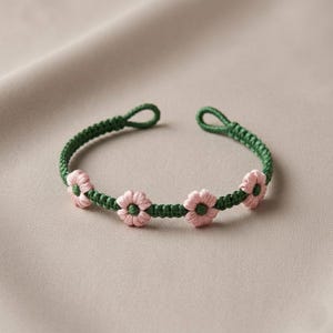 May include: A green macrame bracelet featuring pink flower accents. The bracelet is open-ended with a loop closure. The flowers are evenly spaced along the bracelet's front, creating a delicate and feminine design.