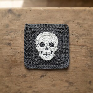 Könnte beinhalten: Ein quadratischer Häkeluntersetzer mit dunkel anthrazitfarbenem Hintergrund und einem mittig platzierten weißen Totenkopfmotiv. Das Stück ist mit einer sichtbaren Garnstruktur und einem gleichmäßigen Stichmuster im gesamten Quadrat gefertigt.