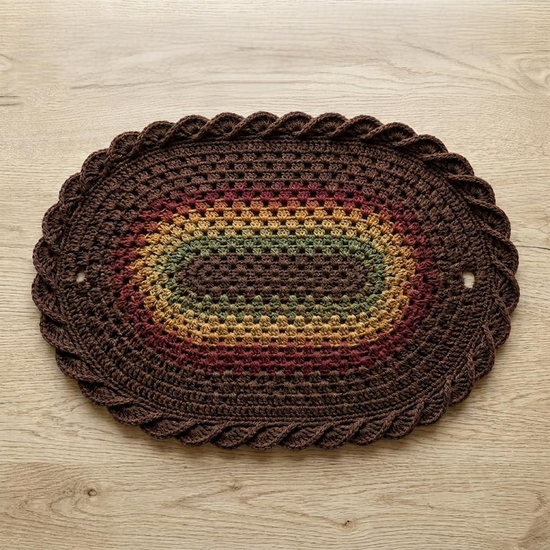 Op de afbeelding: Ovale, handgemaakte gehaakte vloerkleed in bruin, rood, geel en groen. Het kleed heeft een geschulpte rand en een centraal ovaal ontwerp met kleurovergangen. Het kleed ligt op een houten oppervlak.
