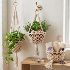 May include: Macrame hanging baskets with plants and herbs. One basket holds a green leafy plant, another contains fresh herbs and alliums, and a third holds rolled towels and soaps. The baskets are suspended from wooden rings and a brass hook.