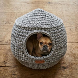 May include: A gray, crocheted dog bed shaped like a dome, with a circular opening. A brown French Bulldog is inside the bed, looking out. The bed has a small, rectangular tag that reads "COZY-N-CO."