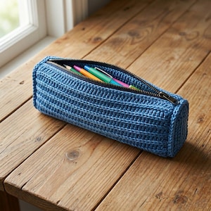 Può includere: Un astuccio blu all'uncinetto con una cerniera nera, parzialmente aperto per rivelare penne e matite colorate. L'astuccio cilindrico è realizzato in un materiale intrecciato e strutturato. L'astuccio è appoggiato su una superficie di legno.