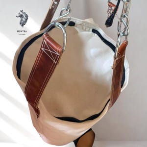 May include: A beige canvas tote bag with brown leather straps and silver metal hardware. The bag has a black trim and is suspended from a chain. The brand logo, "MONTRA LEATHER", is visible in the upper left corner.