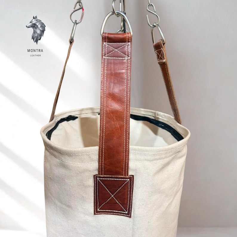May include: A cream canvas bucket bag with brown leather straps and accents, suspended from metal rings and chains. The leather straps are stitched with white thread. The bag has a black trim around the top opening.