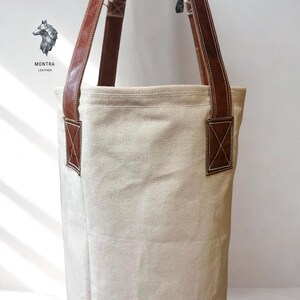 May include: Cream canvas tote bag with brown leather handles and detailing. The bag has rectangular leather patches with an X-shaped stitch on the sides. The brand logo, "MONTRA LEATHER", is visible in the upper left corner.
