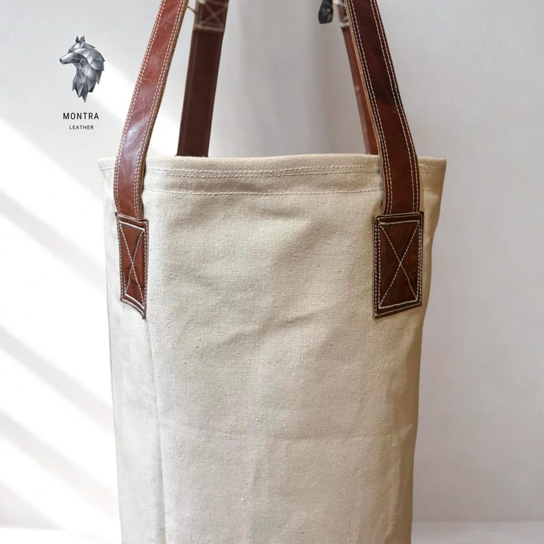 May include: Cream canvas tote bag with brown leather handles and detailing. The bag has rectangular leather patches with an X-shaped stitch on the sides. The brand logo, "MONTRA LEATHER", is visible in the upper left corner.