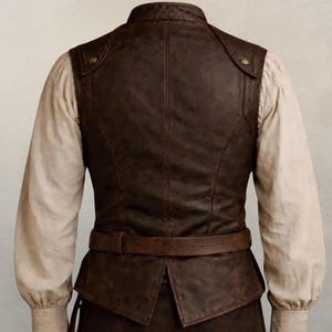 May include: A dark brown leather vest featuring a high collar, structured seams, and a matching leather belt at the waist. The vest is worn over a long-sleeved, off-white shirt with gathered cuffs. The design includes metallic buttons at the shoulders.
