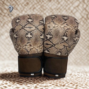 May include: A pair of boxing gloves with a snakeskin pattern in shades of brown and tan. The gloves have dark brown wrist straps and are displayed against a textured background. The brand logo is visible in the upper left corner.