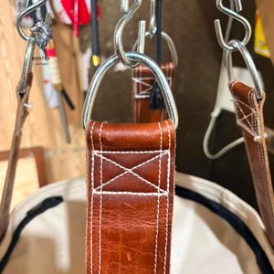 May include: Close-up of a brown leather strap with white stitching, connected to a metal ring. The strap is part of a larger item. The logo "MONTRA LEATHER" is visible in the upper left corner. The leather has a textured appearance.
