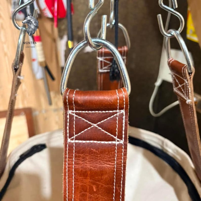 May include: Close-up of a brown leather strap with white stitching, connected to a metal ring. The strap is part of a larger item. The logo "MONTRA LEATHER" is visible in the upper left corner. The leather has a textured appearance.