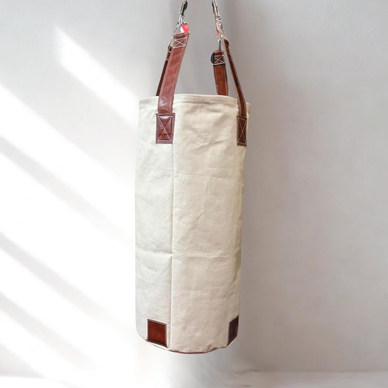 May include: A cream-coloured, cylindrical punch bag with brown leather accents. The bag has brown leather straps and reinforced corners. The bag is suspended from a metal hook.