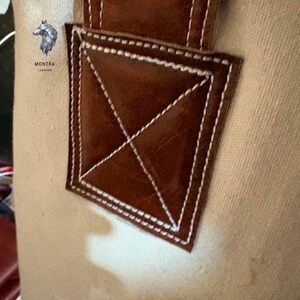 May include: Close-up of a canvas bag with a brown leather accent. The leather is stitched with white thread in a diamond pattern. The bag has a logo with a wolf and the text "MONTRA LEATHER".