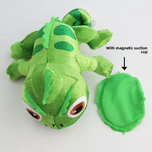 May include: A green plush chameleon toy with brown and white eyes and a segmented body. The toy includes a separate green magnetic suction cup. The text "With magnetic suction cup" is also visible.