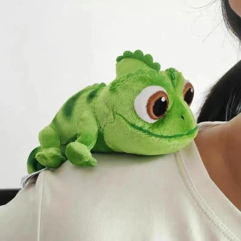 May include: A plush green chameleon toy with large brown and white eyes, and a ridged crest. The toy has darker green stripes and is resting on a white surface.