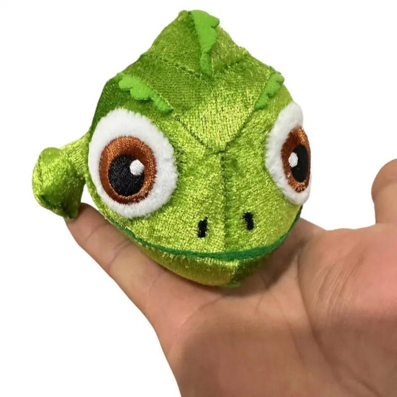 May include: A plush green chameleon toy with large brown and white eyes. The stuffed animal has a textured, velvet-like appearance and is held in a hand. The toy has a triangular head and a small, curved tail.