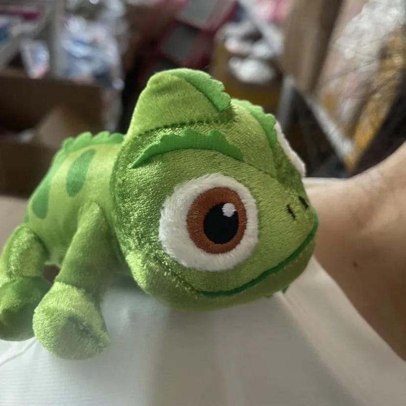 May include: A plush green chameleon toy with a detailed design. The toy has large brown and white eyes, a textured body, and a curled tail. The chameleon has a light green belly and darker green spots.