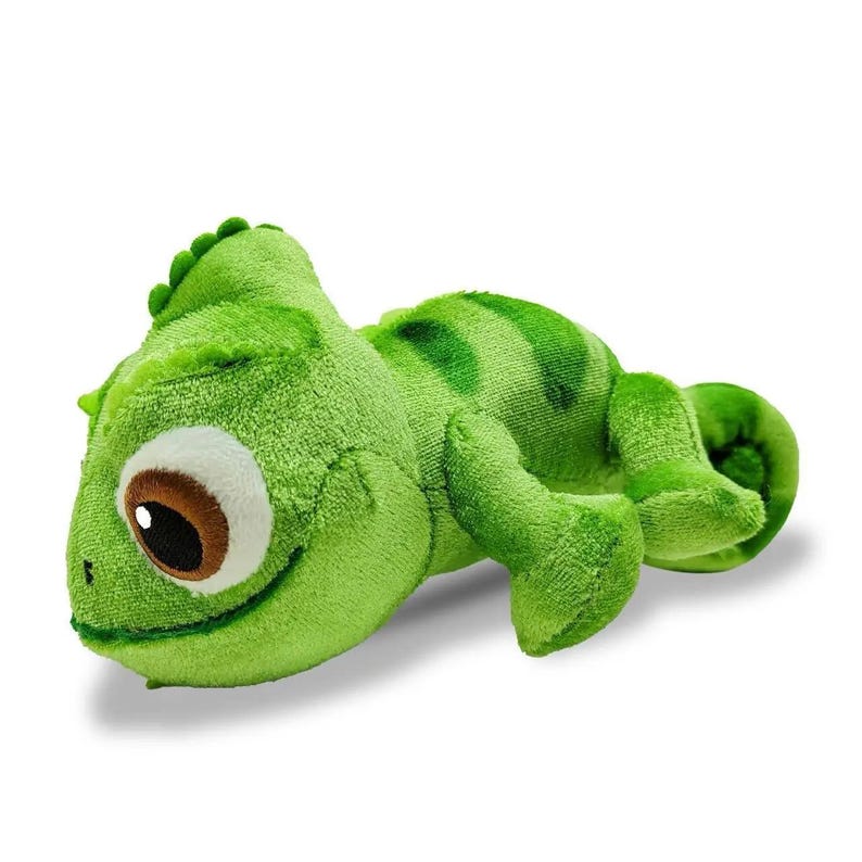 May include: A plush green chameleon toy with a smiling expression. The toy has a textured surface, a brown and white eye, and darker green markings on its back. The tail is curled, and the toy is lying down.