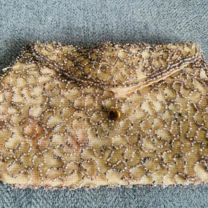 May include: A rectangular clutch bag featuring a light peach fabric base covered in intricate, swirling patterns of metallic silver beads. The bag has a fold-over flap closure secured by a single, small, dark brown circular button at the centre.
