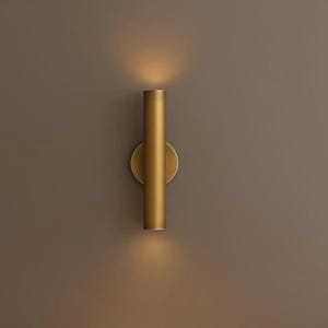 Peut inclure: Une applique murale dorée avec un luminaire cylindrique. L'applique a une base circulaire et émet de la lumière vers le haut et vers le bas. Le luminaire est monté sur un mur marron.