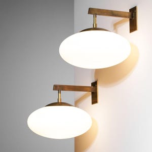 May include: Two wall-mounted sconces with a brass finish and oval, white glass shades. The sconces are mounted on a white wall, casting a warm glow. The design is minimalist and modern, suitable for interior lighting.