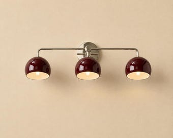 May include: A three-light wall fixture featuring a polished nickel finish metal bar and circular wall mount. Three glossy, deep burgundy dome-shaped shades are attached to the horizontal arm, each housing a single light bulb with a white interior.