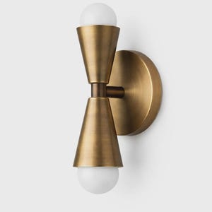 May include: A brass wall sconce featuring a circular backplate and two conical shades. Each shade houses a white, spherical light bulb. The fixture has a warm, metallic finish and a contemporary design.