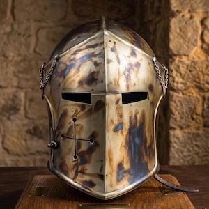 May include: A weathered metal knight's helmet with a cross design on the front. The helmet has a riveted construction, eye slits, and a chin strap. The metal has a mottled, aged appearance with shades of gold, blue, and brown.
