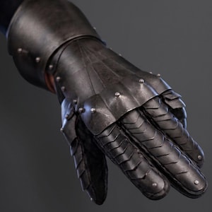 May include: Close-up of a black metal gauntlet, part of a medieval suit of armour. The gauntlet features articulated plates and rivets, designed to protect the hand and wrist. The armour is dark grey and has a slightly weathered appearance.