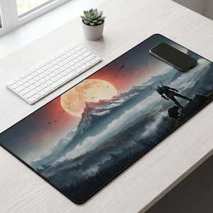 May include: A black rectangular desk mat featuring a mountain landscape with a large orange moon and a silhouetted figure. A white keyboard and a black smartphone are also on the desk.