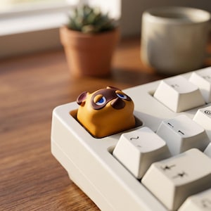 May include: A custom mechanical keyboard keycap shaped like the head of a tanuki character with brown ears, a dark brown mask, and blue eyes, installed on a white keyboard among standard white keys with black lettering.